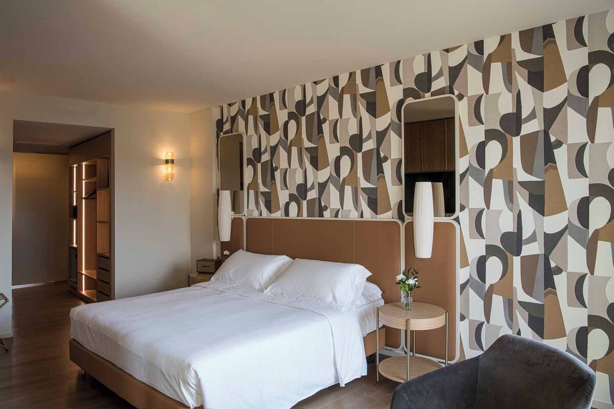 A hotel room with a large bed, a bedside table with a vase, and a mirror on the wall.