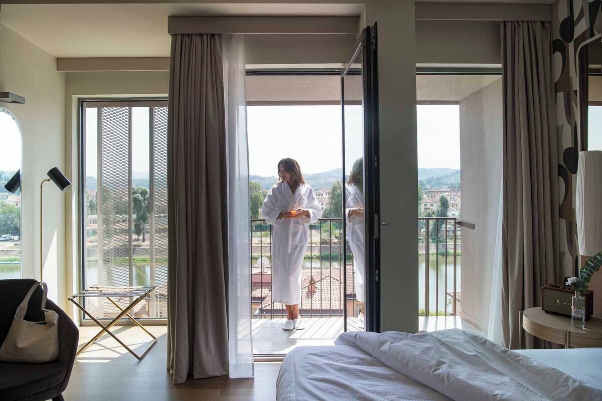 A hotel room with a bed, a chair, a table with a radio, and a balcony with a view of the countryside.