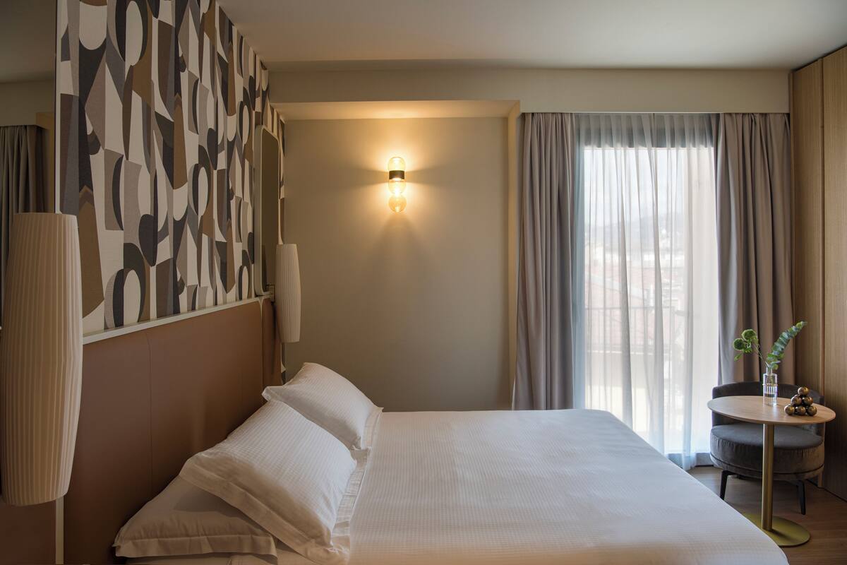 A hotel room with a large bed, a bedside table with a plant, a wall-mounted light, and a view of the city through the window.