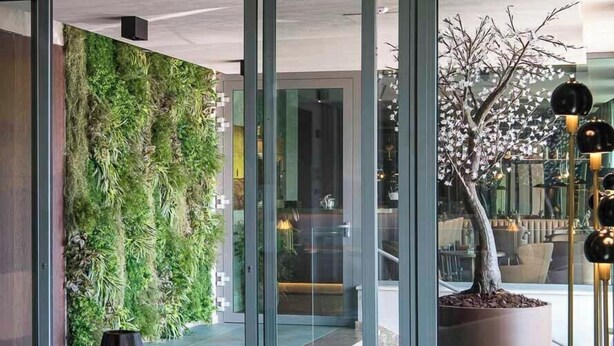 A modern interior with a curved sofa, two tall vases, a green vertical garden, and a potted tree. - Columbus Urban Resort (Florence)