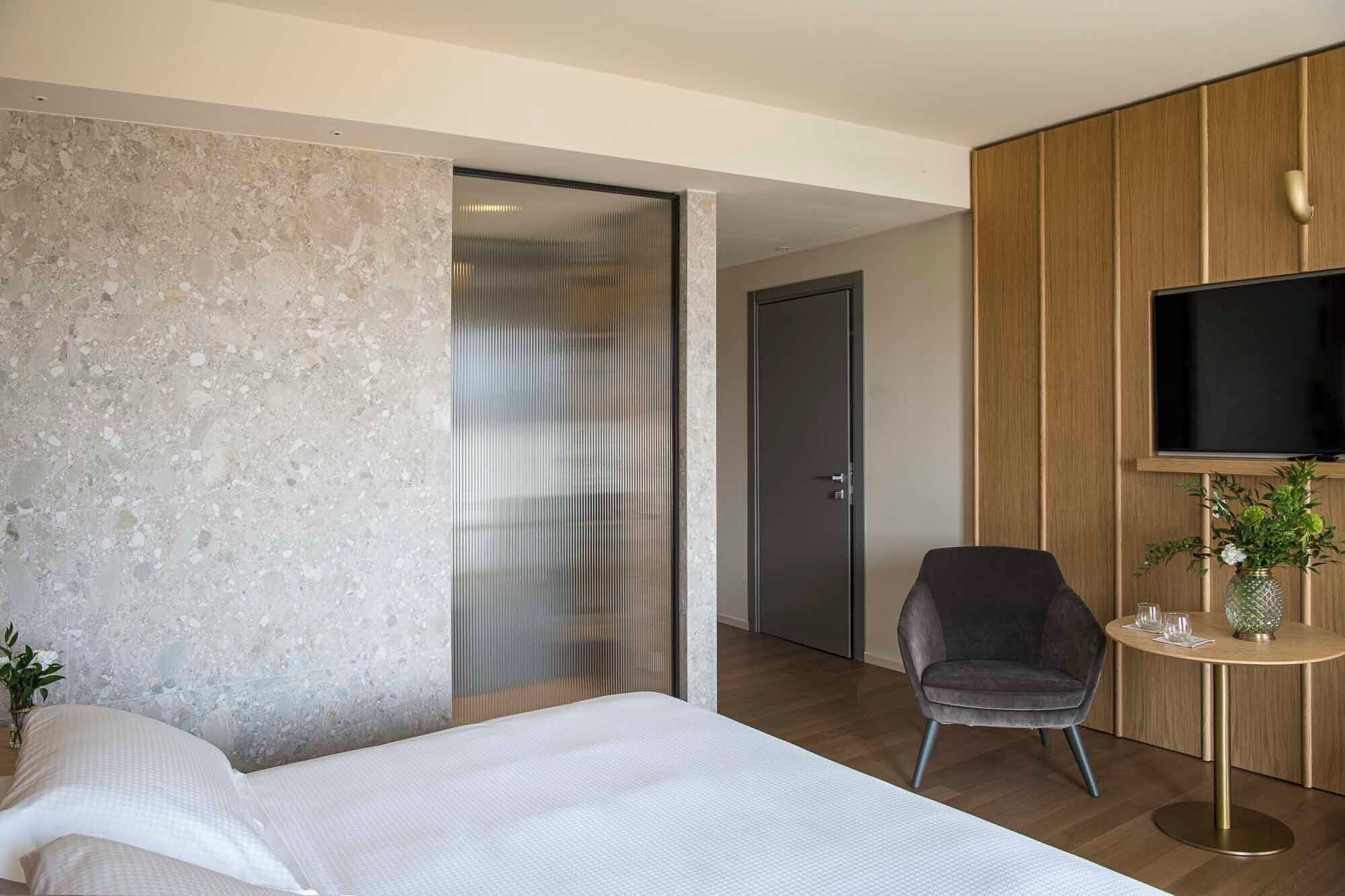 Una habitación de hotel moderna con una cama grande, un área de descanso con una silla, una mesita con un florero y un televisor montado en un panel de madera.