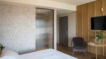 Una habitación de hotel moderna con una cama grande, un área de descanso con una silla, una mesita con un florero y un televisor montado en un panel de madera.
