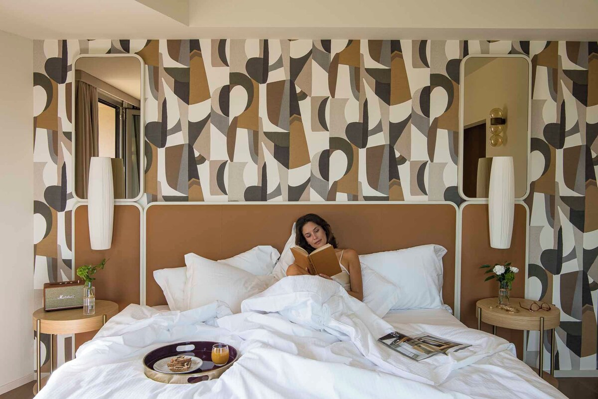 A hotel room with a modern abstract wall design, a large bed with white bedding, a bedside table with a radio, and a tray with food and a drink.