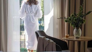 A hotel room with a bed, a chair, a table with a vase and a glass of water, and a view of greenery outside. - Columbus Urban Resort (Florence)