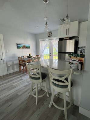A modern kitchen with white cabinetry, a stainless steel refrigerator, and a dining area with two white chairs and a table.