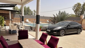 A covered outdoor seating area with tables and chairs, a pool, and a black car parked nearby.