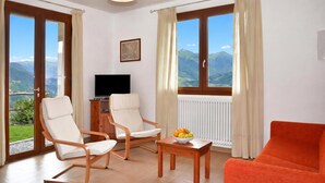 Una habitación con dos sillas, una mesa con fruta, un televisor y ventanas grandes con cortinas.