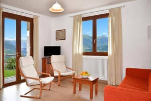 A room with two chairs, a table with fruit, a television, and large windows with curtains. - Costa Del Fuoco (Bellano)