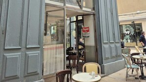 Ein Café mit einer Schrifttafel, auf der "CAFE DE L'HORLOGE" steht, und Außenplätzen mit Tischen und Stühlen.