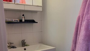 A bathroom with a white sink, a mirror, a shelf with toiletries, and a pink towel hanging on the wall.
