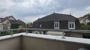 A balcony with a wooden table and a woven stool, overlooking a residential area with houses and a cloudy sky.