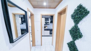 A hallway with a kitchen area, white tiled flooring, wooden doors, and a mirror on the left.