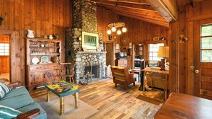 Interior rústico de cabana, com paredes de madeira, lareira de pedra, mesa de jantar de madeira e um espaço de estar acolhedor.