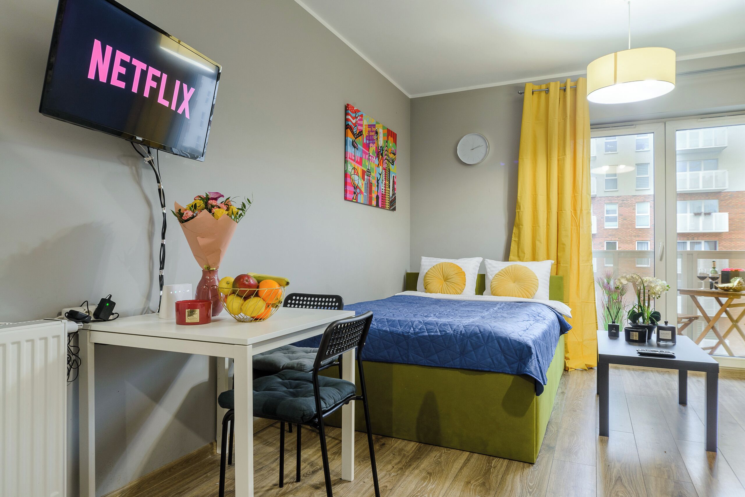 Una habitación de hotel moderna con una cama, un escritorio con una silla, una mesa con un jarrón de flores, un reloj en la pared y una ventana con cortinas amarillas.