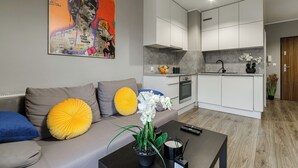 A modern living room with a grey sofa, yellow and blue cushions, a coffee table with a potted orchid, and a kitchen area with white cabinets and a grey backsplash. - Lumina modern studio with balcony (Lodz)