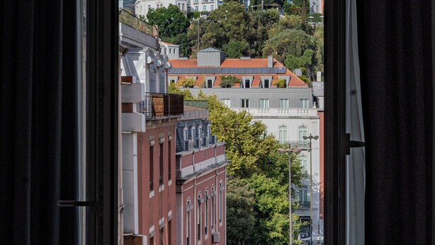 Vista desde una ventana con cortinas, que muestra un paisaje urbano con edificios y una colina.