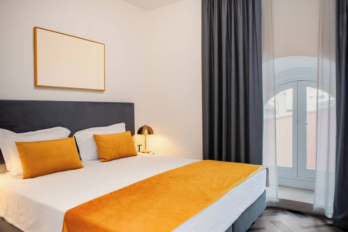 A hotel room with a bed, a bedside table with a lamp, a framed picture on the wall, and a view through a window with curtains.