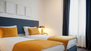 Habitación de hotel con dos camas, cada una con cabecero azul y ropa de cama blanca. Hay una lámpara sobre una mesita de noche y una ventana con cortinas.