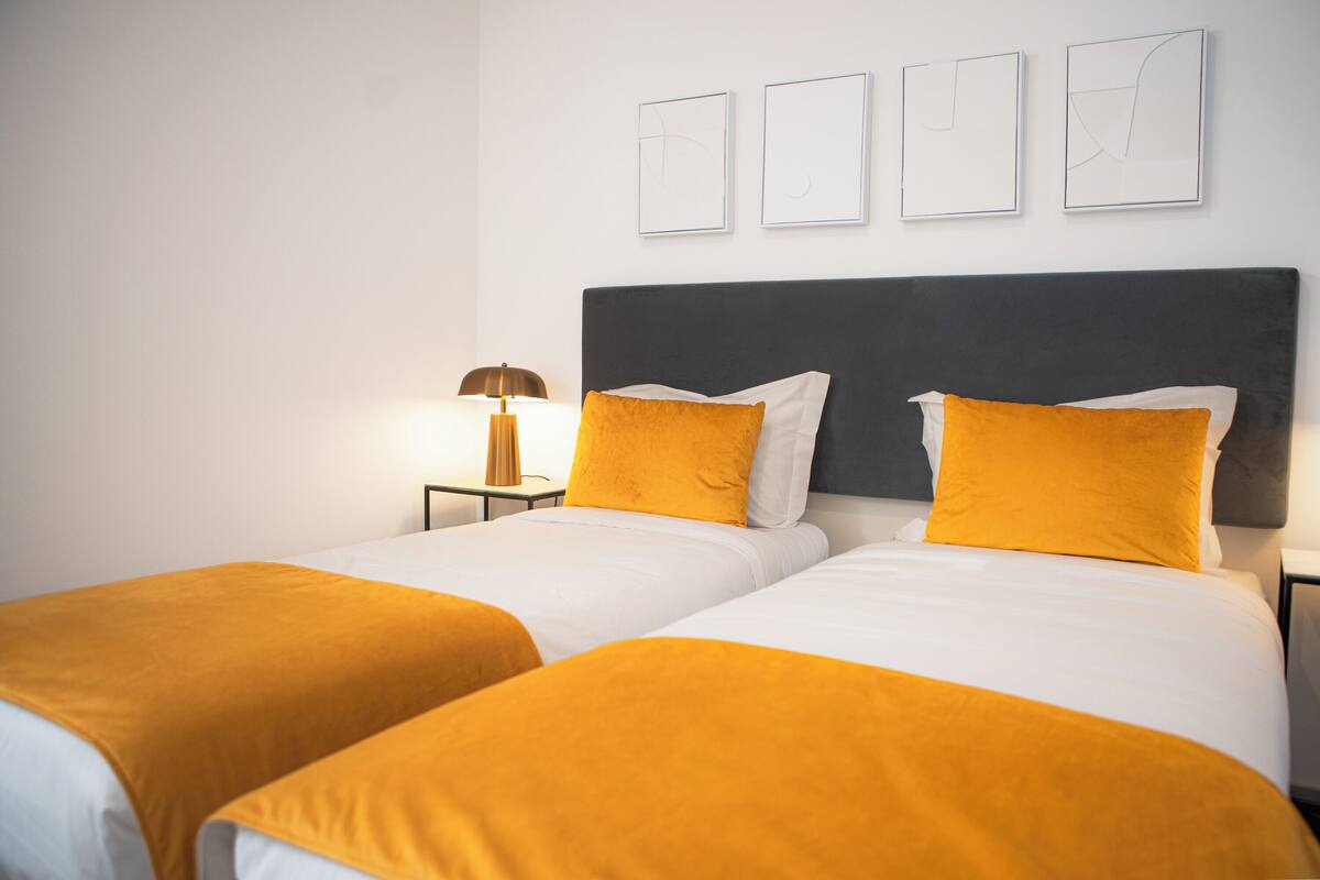 A hotel room with two beds, each with a white and orange bedspread, a dark headboard, and a nightstand with a lamp.