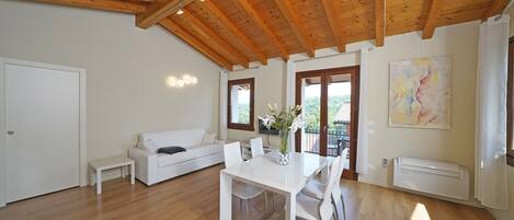 A spacious living area with a wooden ceiling, a white sofa, a dining table with chairs, and a large window with a view of greenery.