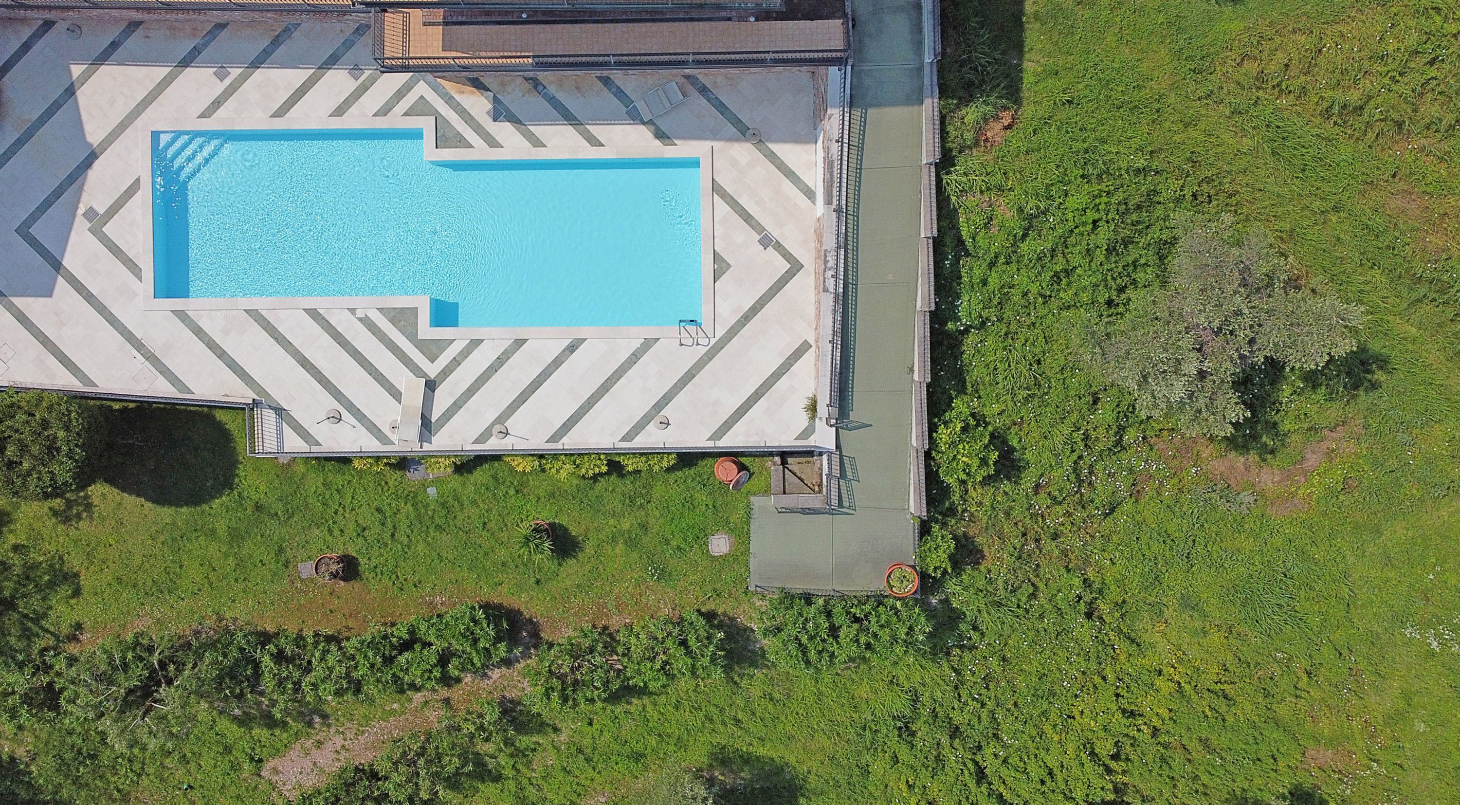 An aerial view of a swimming pool with a geometric tile pattern, surrounded by a grassy area with some shrubs.