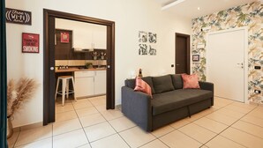 Un salon moderne avec un canapé gris, un coin cuisine doté d’un bar et une porte recouverte d’un papier peint aux motifs tropicaux.