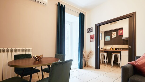 Un salon moderne avec un coin repas, comprenant une table ronde, des chaises vert foncé et un canapé agrémenté d’un coussin rouge.