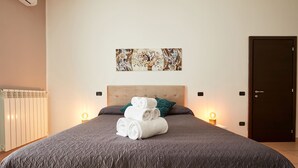 Un lit bien fait, avec des serviettes blanches pliées en rouleau, une couverture de couleur gris foncé et un coussin turquoise.