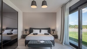 Un dormitorio moderno con una cama grande, dos mesitas de noche, un banco y vistas a un paisaje verde a través de amplias ventanas.