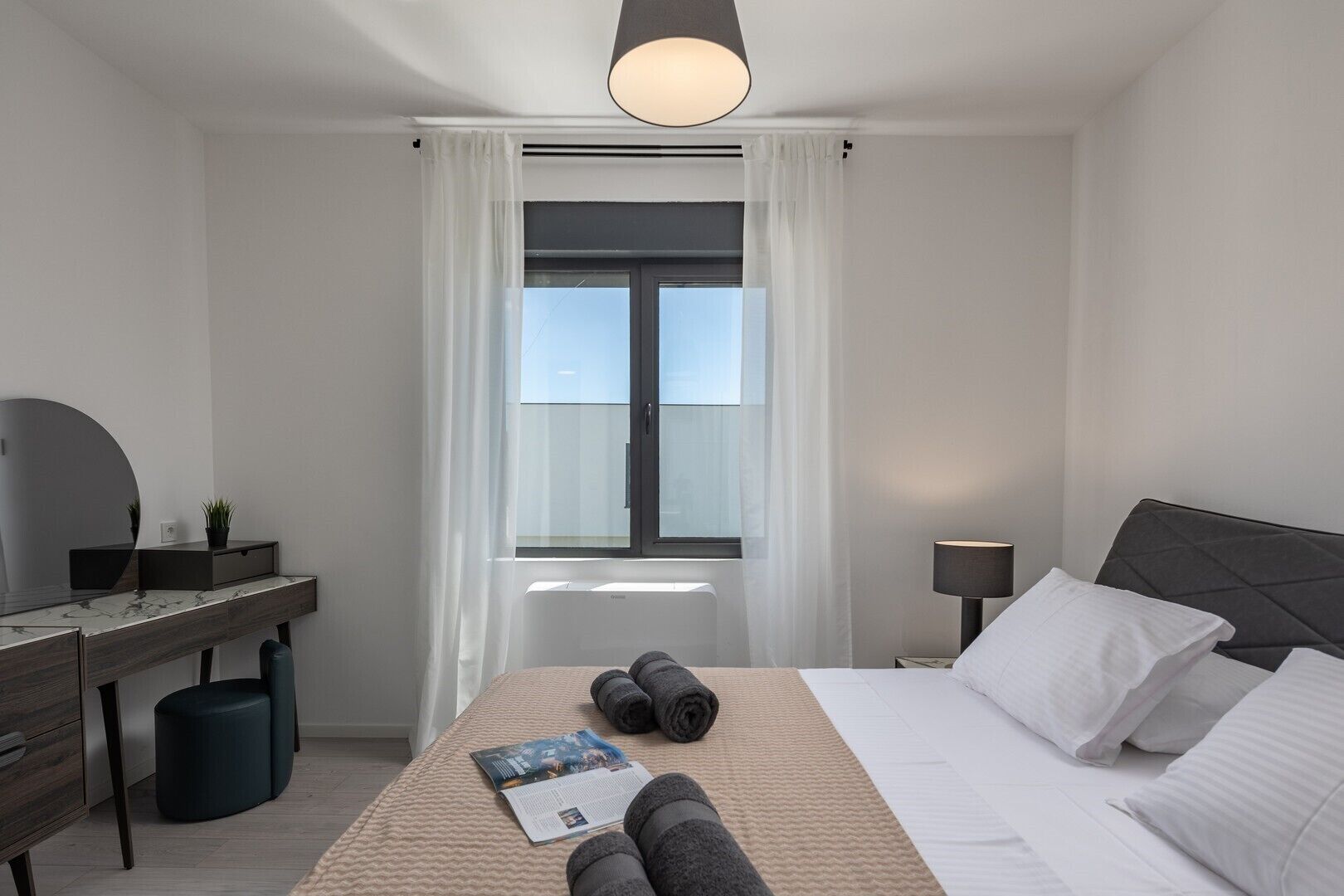 A modern hotel room with a bed, bedside lamp, and a desk with a mirror and small plant.