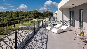 A modern outdoor balcony with white lounge chairs and a wicker chair, overlooking a landscaped garden and a clear blue sky.