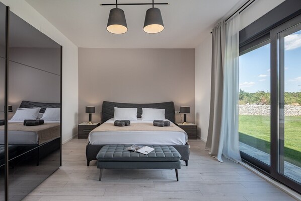 A modern bedroom with a large bed, two bedside tables, a bench, and a view of a green landscape through large windows.