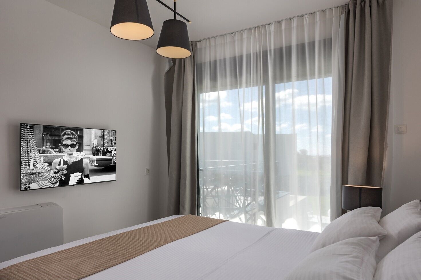 Una habitación de hotel moderna con una cama grande, un televisor montado en la pared y un balcón con cortinas.