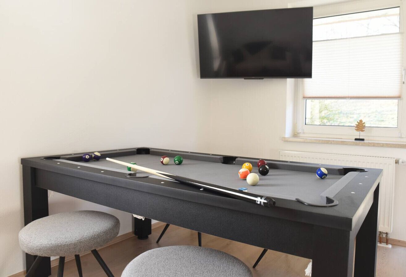 Une table de billard avec des billes et des queues, un téléviseur au-dessus, et une fenêtre avec des stores.