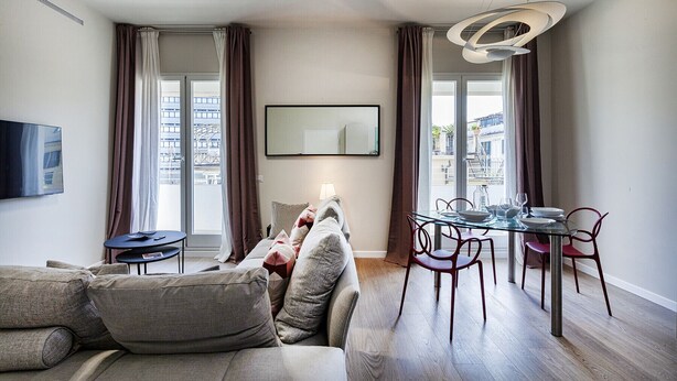 Een moderne woonkamer met een bank, een eettafel met stoelen, een televisie en een groot raam met gordijnen.