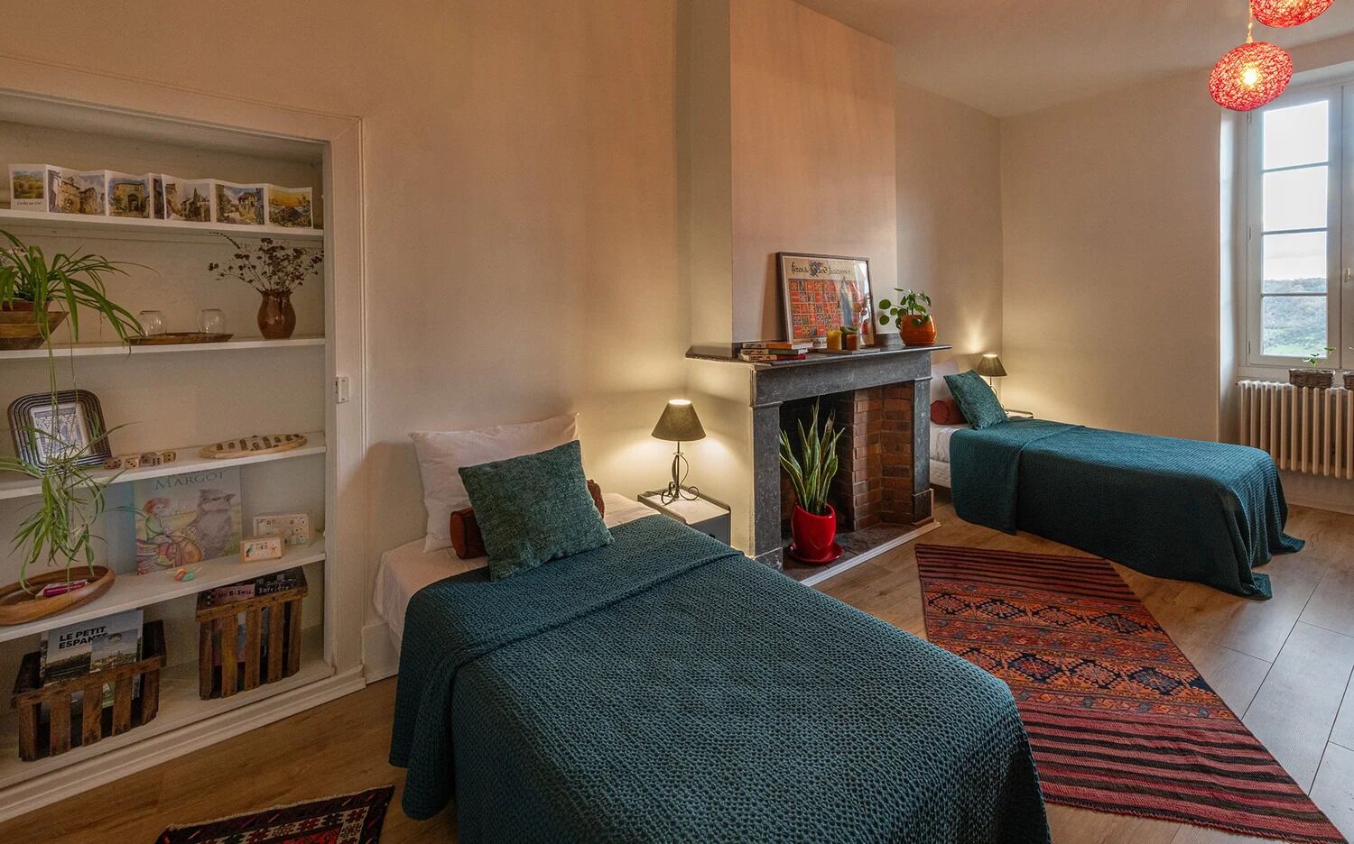 A room with two beds, a fireplace, a bookshelf, and a window with a view.