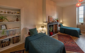 A room with two beds, a fireplace, a bookshelf, and a window with a view.