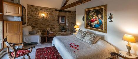 Un dormitorio con pared de piedra, cama con colcha blanca, alfombra roja estampada y un cuadro en la pared.