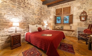 A room with a stone wall, a bed with a red bedspread, a wooden nightstand, a chair, and a window with stained glass.
