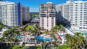 A resort with multiple high-rise buildings, a central swimming pool surrounded by palm trees, and a well-maintained lawn area. - Oceanfront 2BR at The Alexander · Pools + Beach (Miami)
