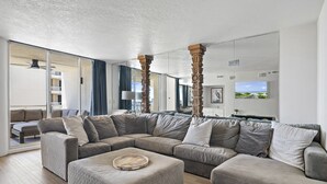 A modern living room with a sectional sofa, ottoman, and a large mirror reflecting the room. - 3BR Harbor front + Panoramic Views + Pool & Beach Access | East Pass Towers (Destin)