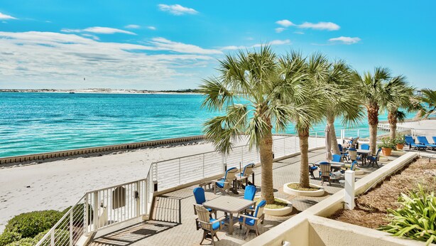 A beachside patio with tables and chairs, palm trees, and a view of the sea. - 3BR Harbor front + Panoramic Views + Pool & Beach Access | East Pass Towers (Destin)