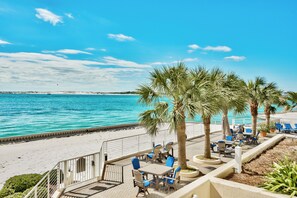 A beachside patio with tables and chairs, palm trees, and a view of the sea. - 3BR Harbor front + Panoramic Views + Pool & Beach Access | East Pass Towers (Destin)