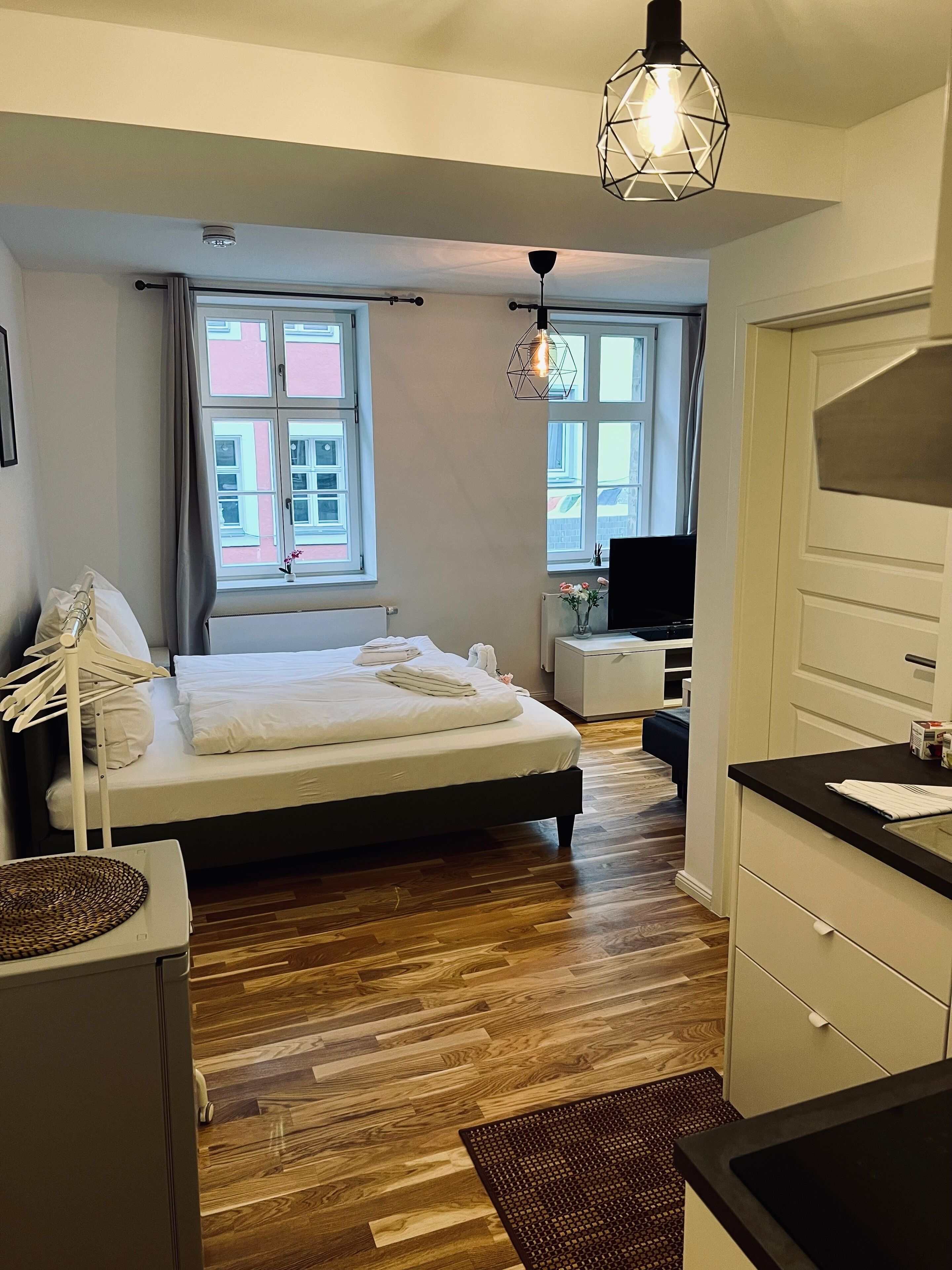 A modern bedroom with a wooden floor, a bed with white linens, a desk with a computer, and a view of buildings through the window.
