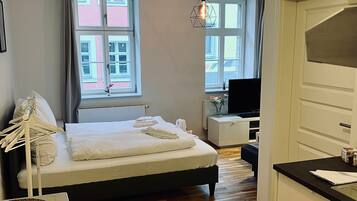 A modern bedroom with a wooden floor, a bed with white linens, a desk with a computer, and a view of buildings through the window.
