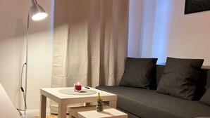 A cozy living room with a grey sofa, a small white table with a candle, a floor lamp, and a peacock feather artwork on the wall. - Central charming apartment in the old town center (Bayreuth)