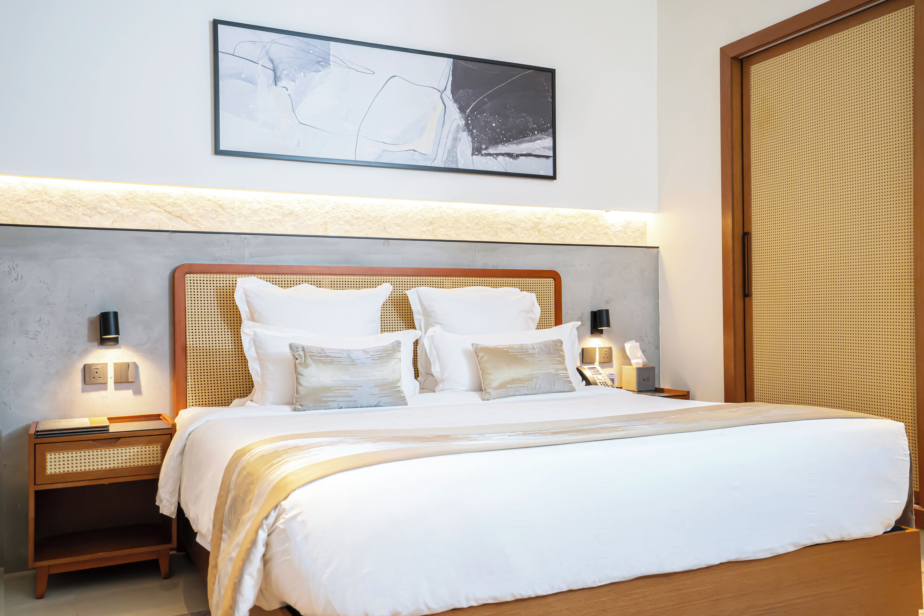 A neatly made bed with white linens and multiple pillows, a bedside table with a lamp, and a wall-mounted abstract painting.
