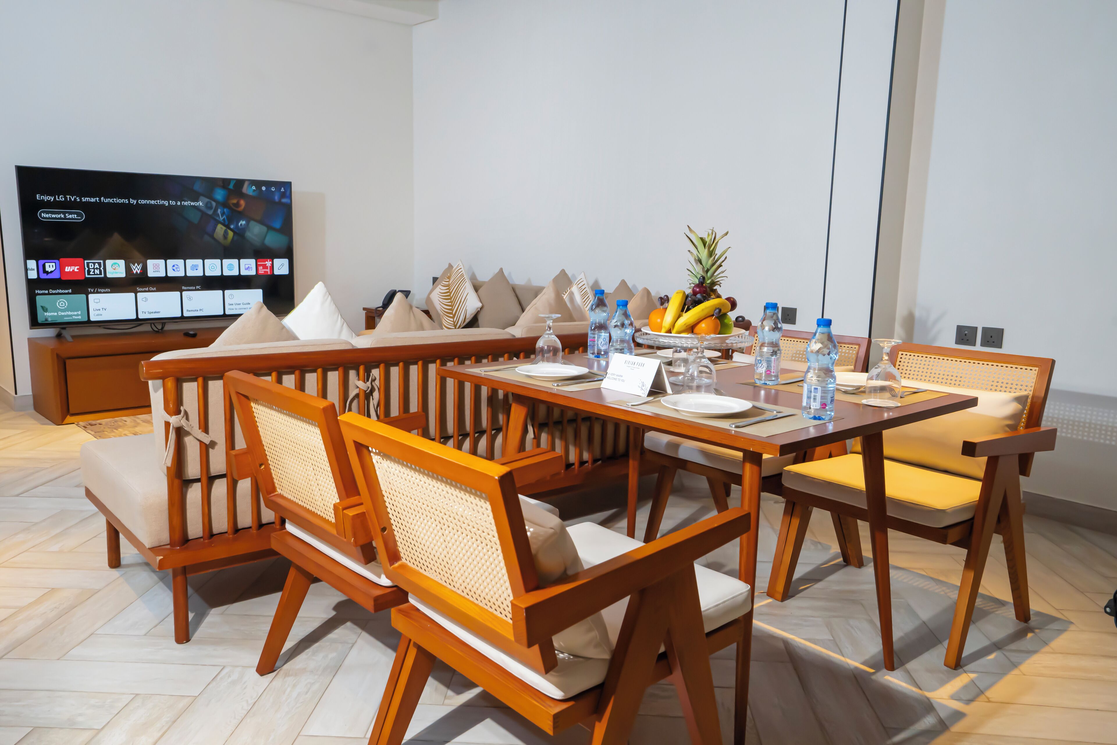 Una sala de estar moderna con una mesa de comedor de madera dispuesta para seis personas, rodeada de sillas de ratán. Un televisor de pantalla plana está montado en la pared, mostrando un panel de control doméstico.