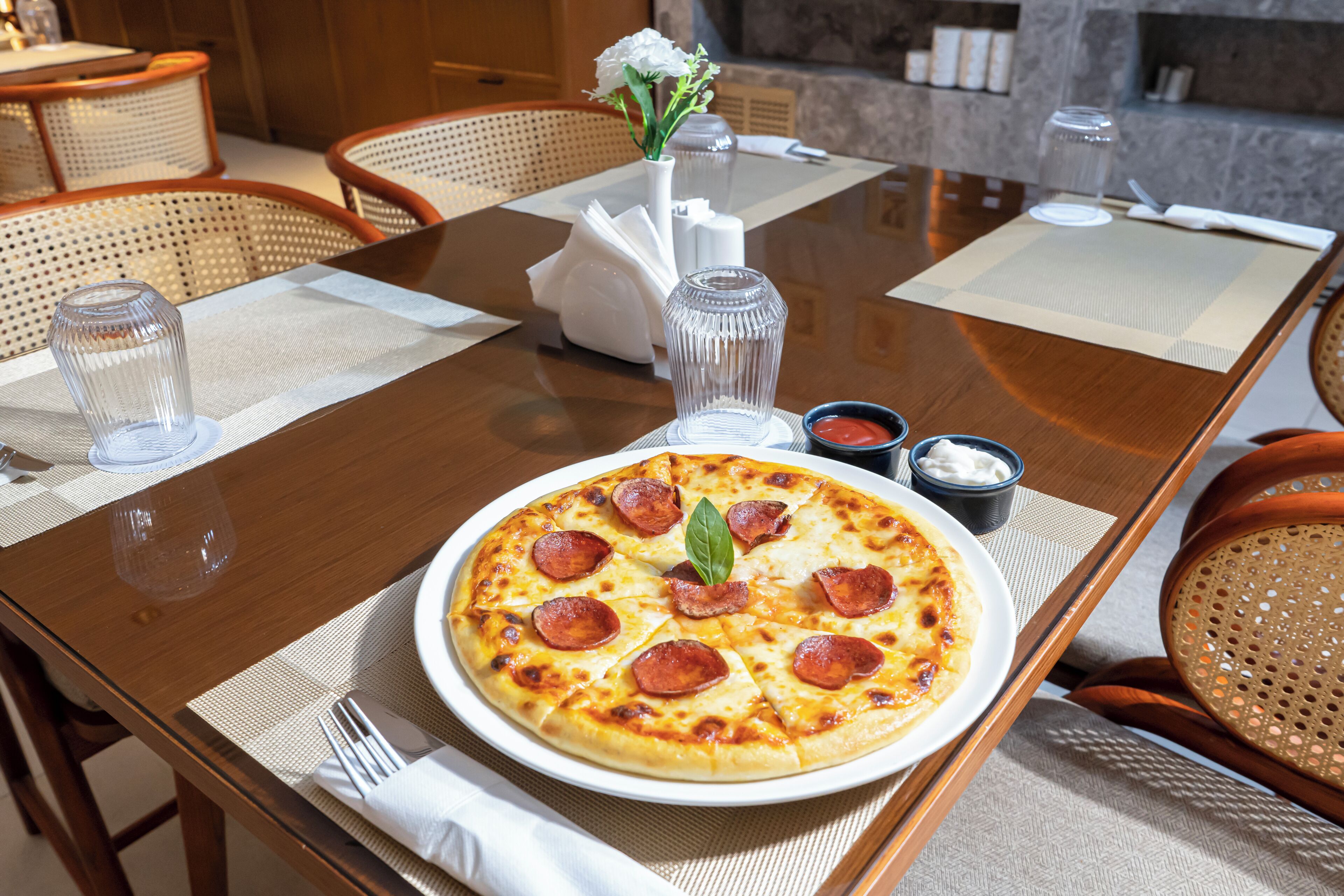 A dining table with a pizza, napkins, cutlery, and a vase with flowers.