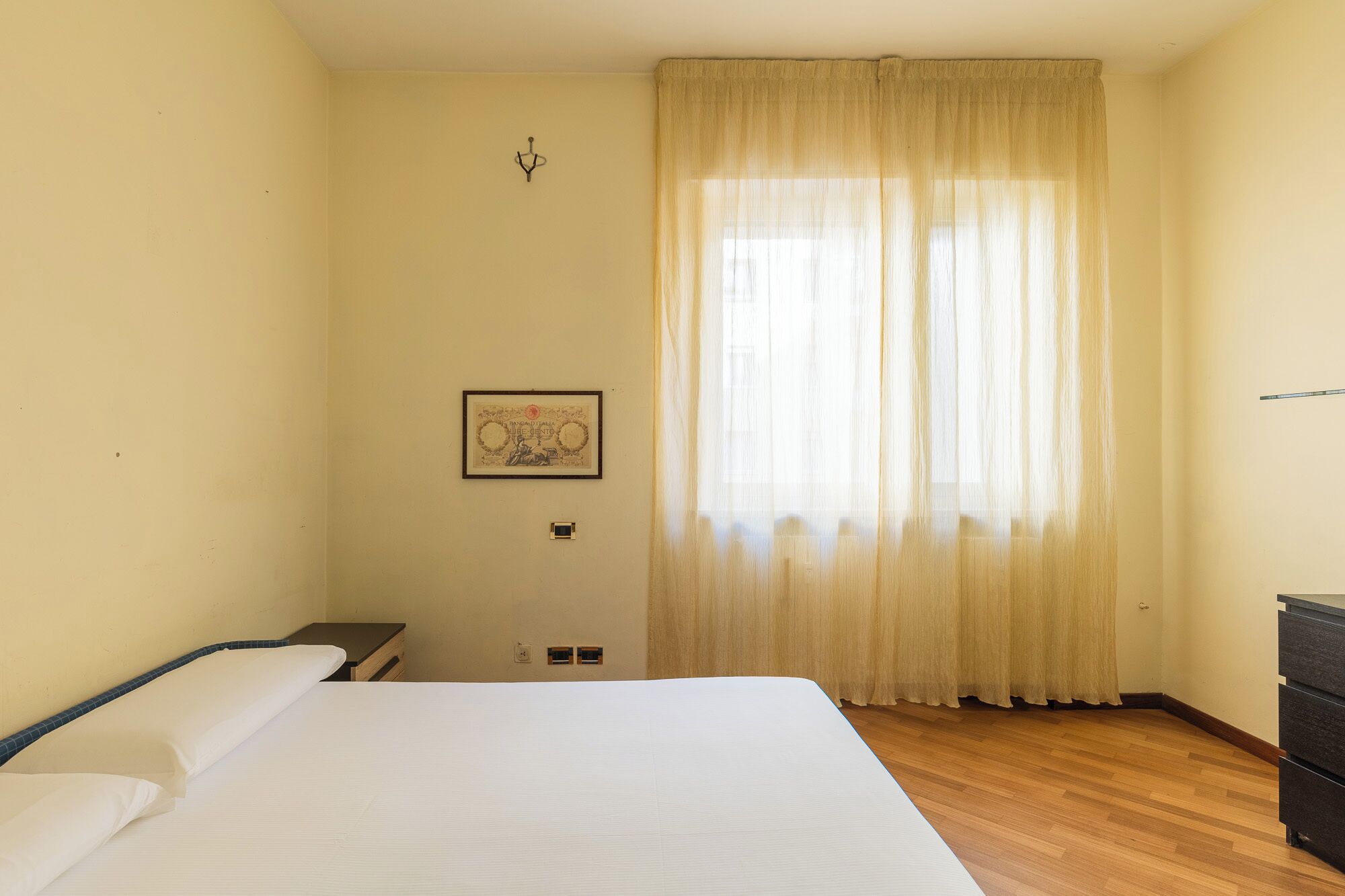 A hotel room with a large bed, a nightstand, a dresser, a window with sheer curtains, and a framed picture on the wall.